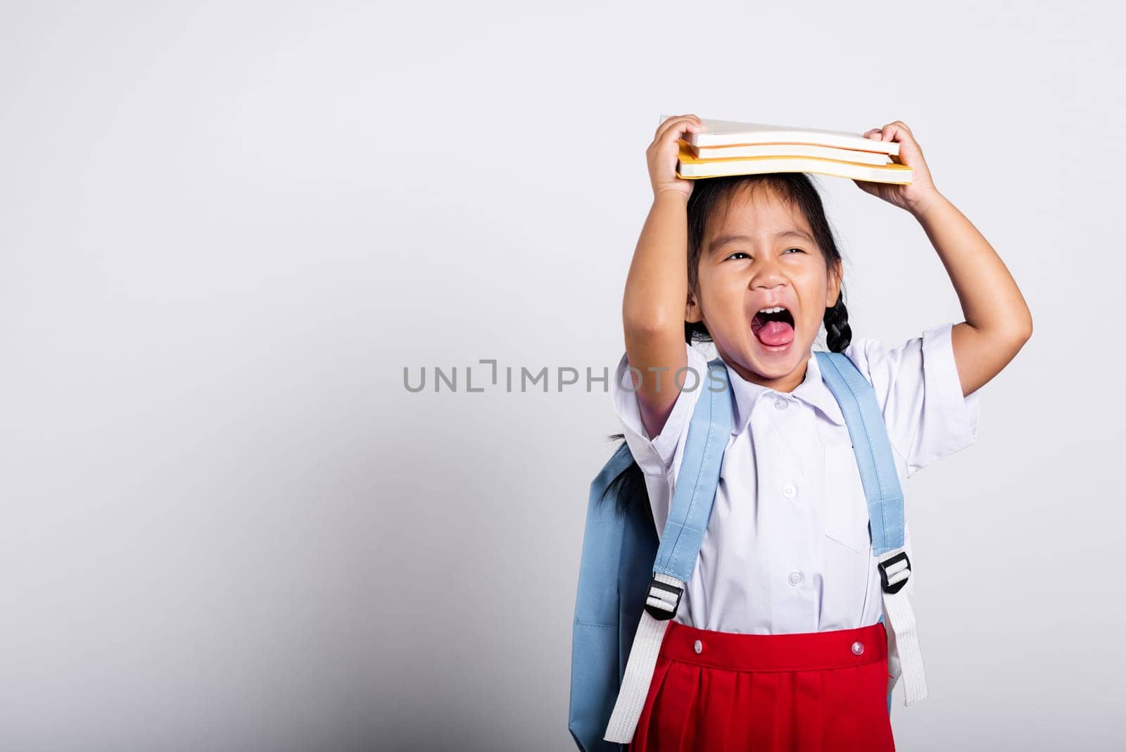 Asian adorable toddler smiling happy wear student thai uniform red skirt stand holding book over head and screaming in studio shot isolated on white background, Portrait little children girl preschool