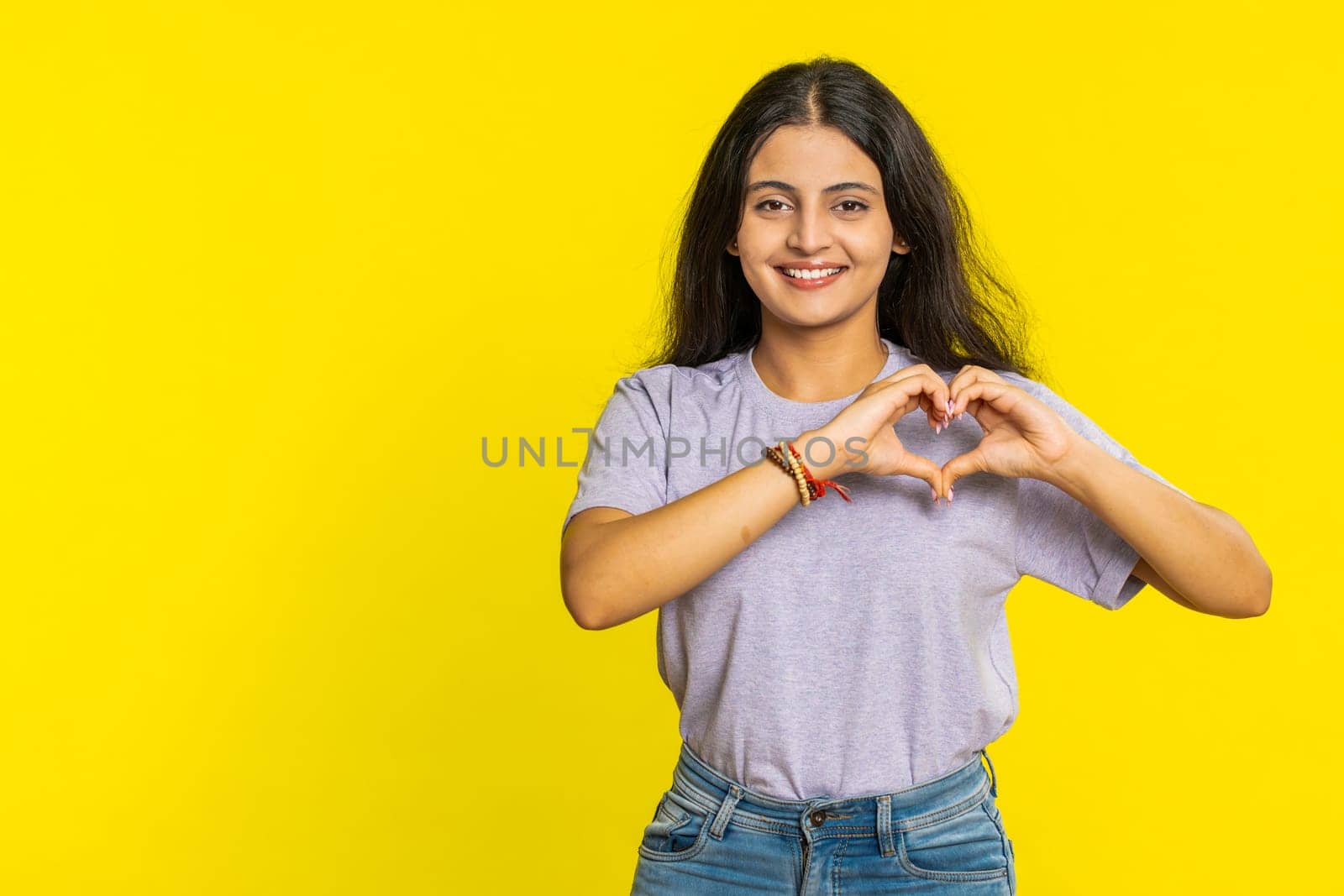 Woman in love. Smiling attractive Indian woman makes heart gesture demonstrates love sign expresses good positive feelings and sympathy. Arabian Hindu young girl isolated on yellow wall background