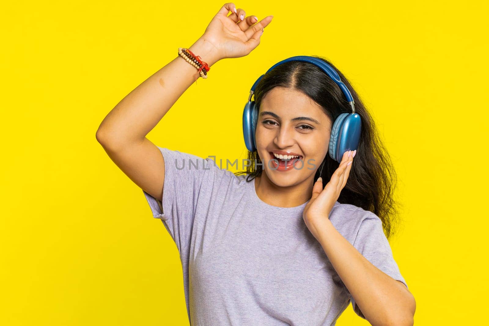 Happy smiling Indian woman listening music in headphones dancing disco fooling around having fun expressive gesticulating hands relaxing chill making funny moves. Hindu girl on yellow background