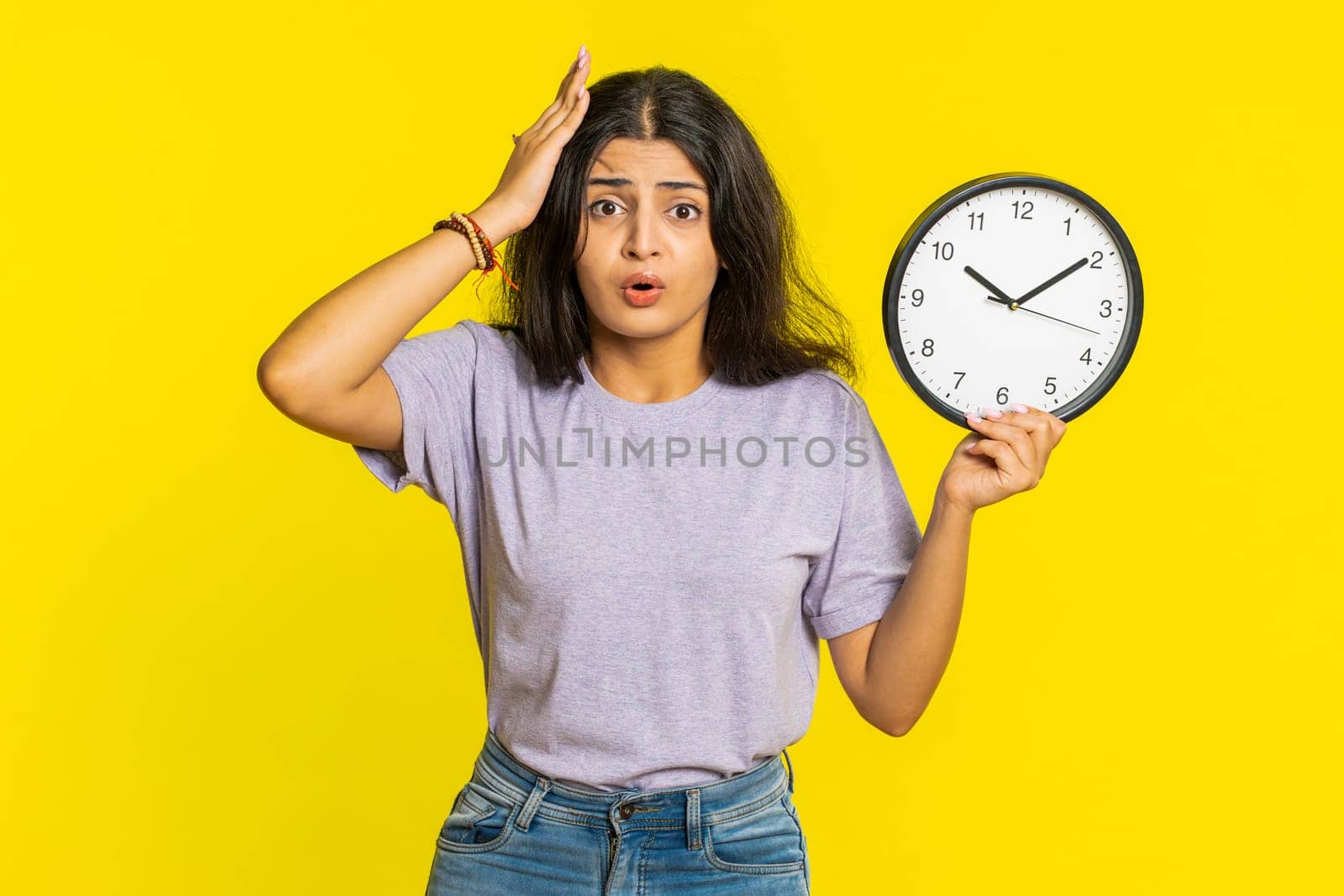 Indian young woman with anxiety checking time on clock, running late to work, being in delay, deadline. Arabian girl looking at hour, minutes, worrying to be punctual isolated on yellow background