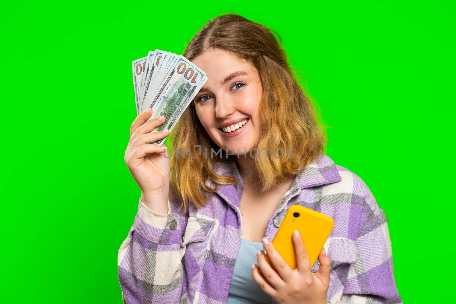 Woman looking smartphone display sincerely rejoicing win, receiving money dollar cash banknotes, celebrating success victory play lottery game. Luck rich girl isolated on green chroma key background