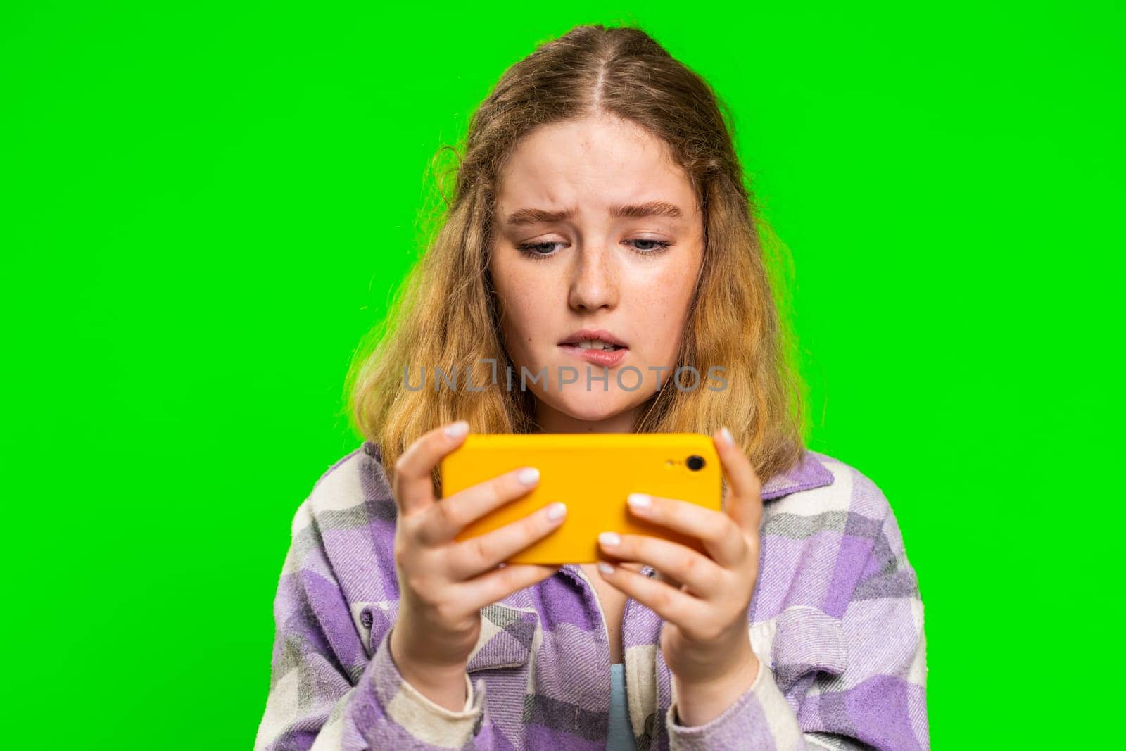 Worried young caucasian woman enthusiastically playing racing drive simulator or shooter video games on smartphone. Happy redhead girl player celebrate victory isolated on green chroma key background