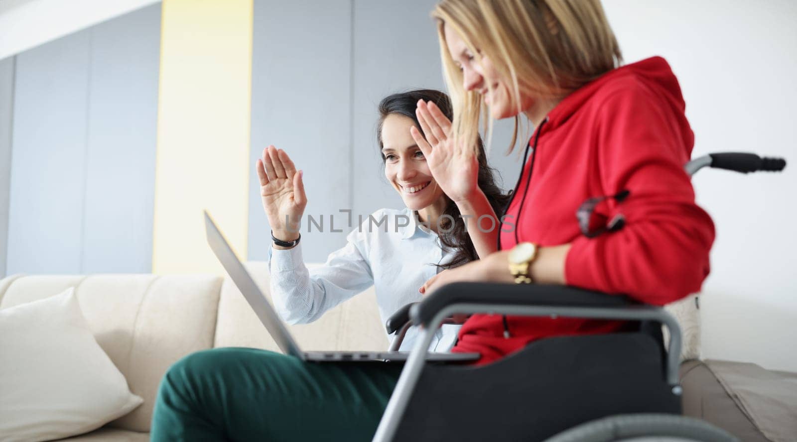 Woman with friend sitting in wheelchair and holding laptop waving to monitor. Remote work for people with disabilities concept