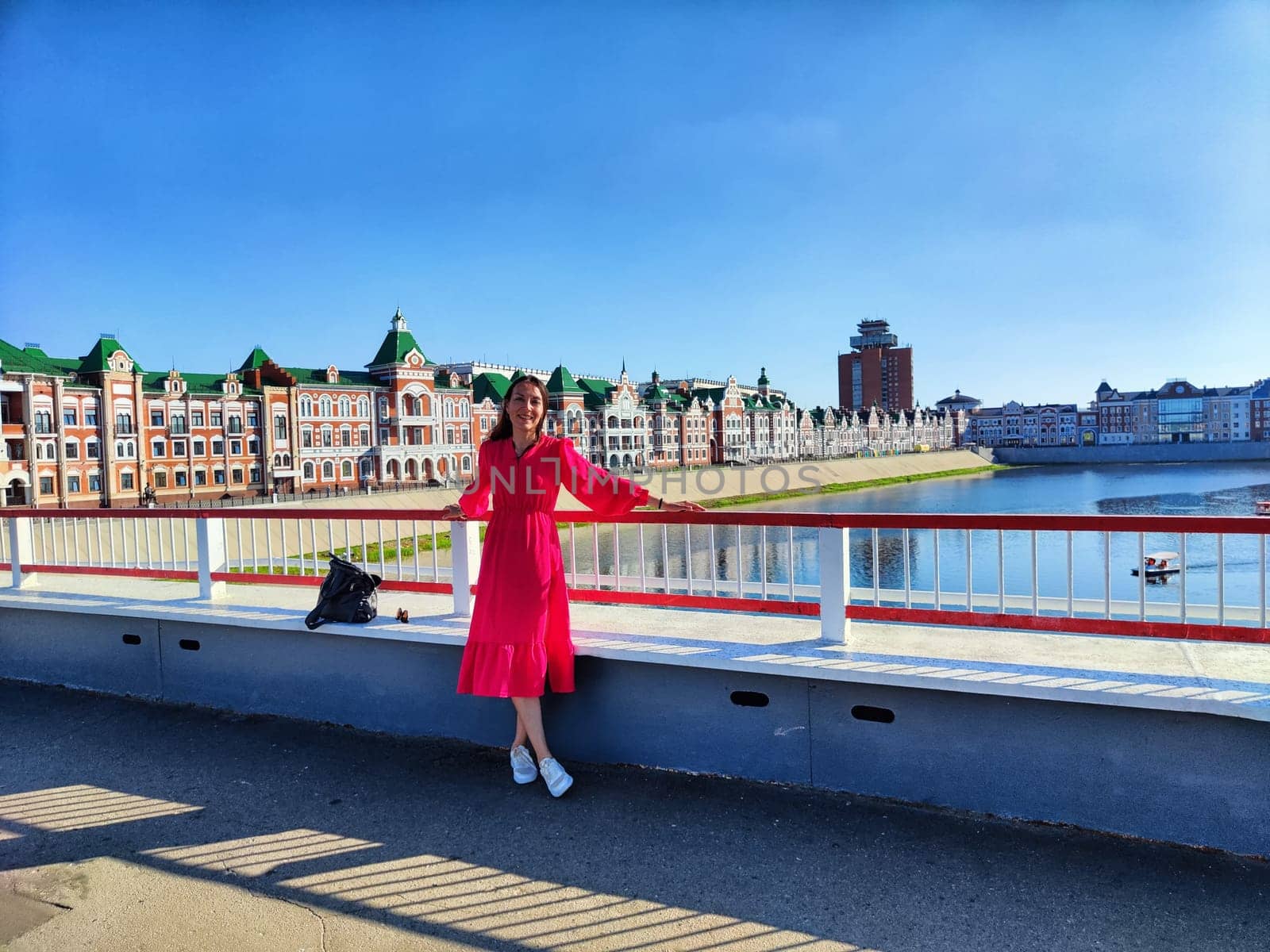 A woman smiles joyfully while standing on a concrete ledge near a river, showcasing vibrant architecture and clear blue sky.