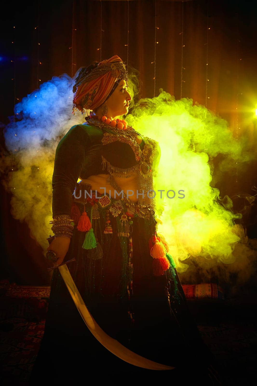 A woman in a colorful headscarf and jewelry poses With a saber in front of wall of colorful tassels and smoke around