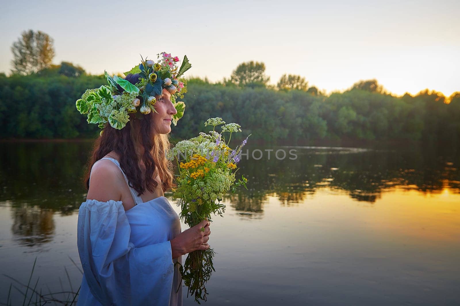 Slavic plump chubby girl in long white dress on the feast of Ivan Kupala with flowers and water in a river or lake on summer evening