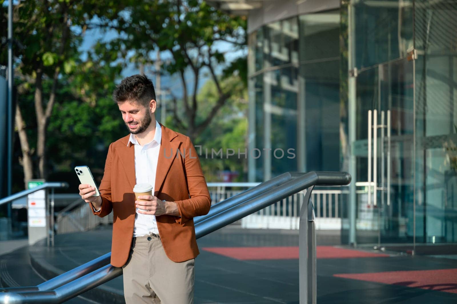 Caucasian businessman holding smartphone and a takeaway coffee cup standing outside office building.