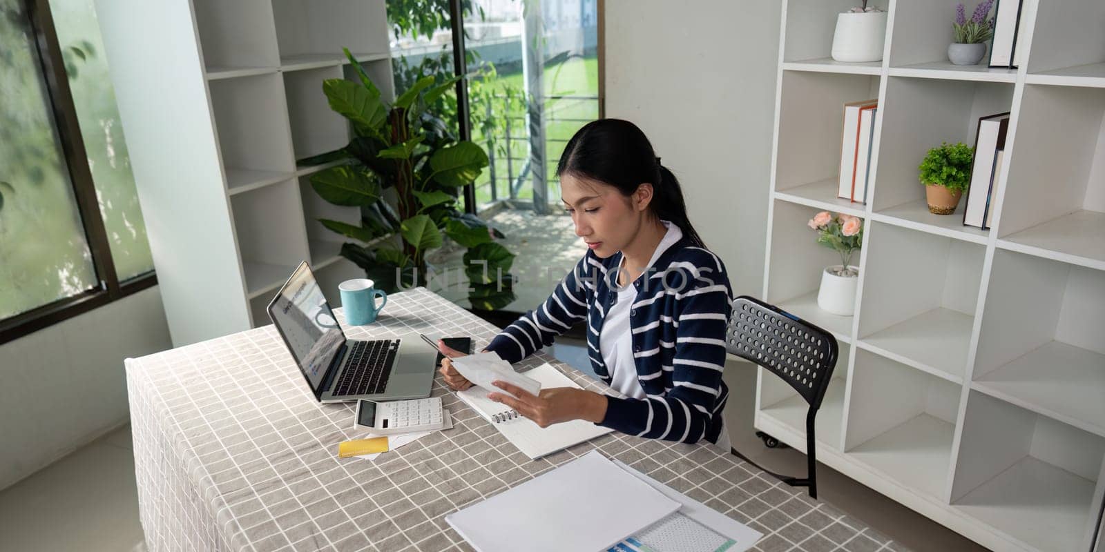 A focused woman calculates her expenses and manages bills at her home workspace, using a laptop and calculator for effective budgeting.