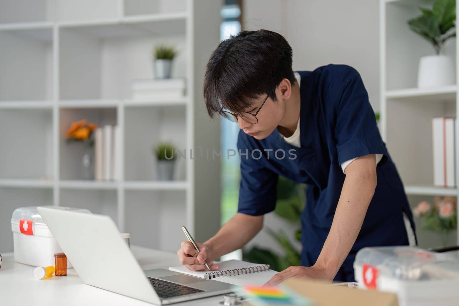 A committed medical student in scrubs writes notes while studying at a desk with a laptop, emphasizing the dedication to learning in healthcare.