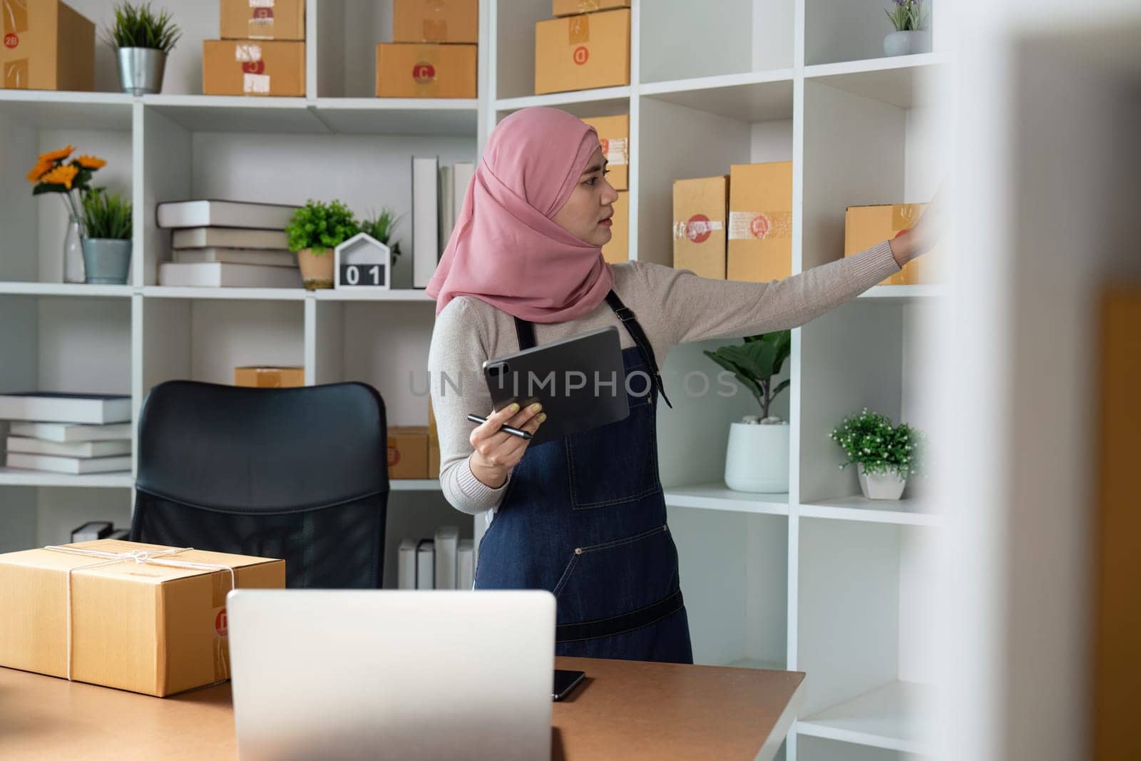 A Muslim entrepreneur in a hijab organizes online orders and manages her business using a tablet in a stylish office setting.
