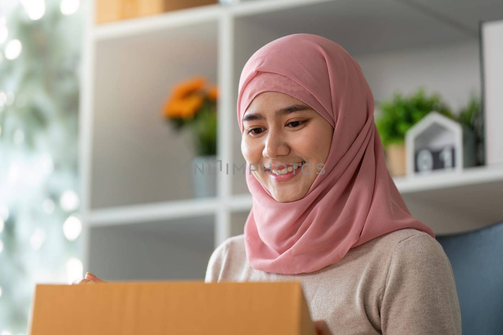 A Muslim woman in a pink hijab smiles as she opens a package, reflecting her success in managing an online business.