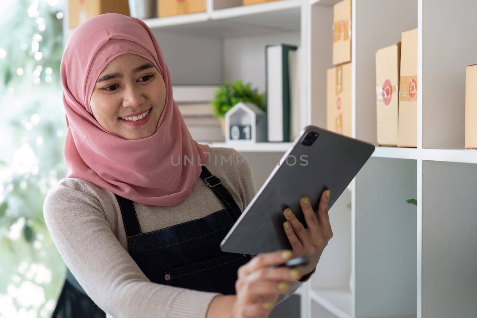 A Muslim woman in a hijab uses a tablet to manage her online sales and inventory, showcasing entrepreneurship and digital commerce.