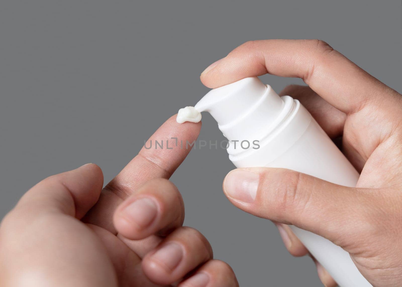 Close up of woman squeezing hand cream on her finger and holding a cosmetic bottle in other hand against grey. Beauty treatment or handcare procedure in a spa salon