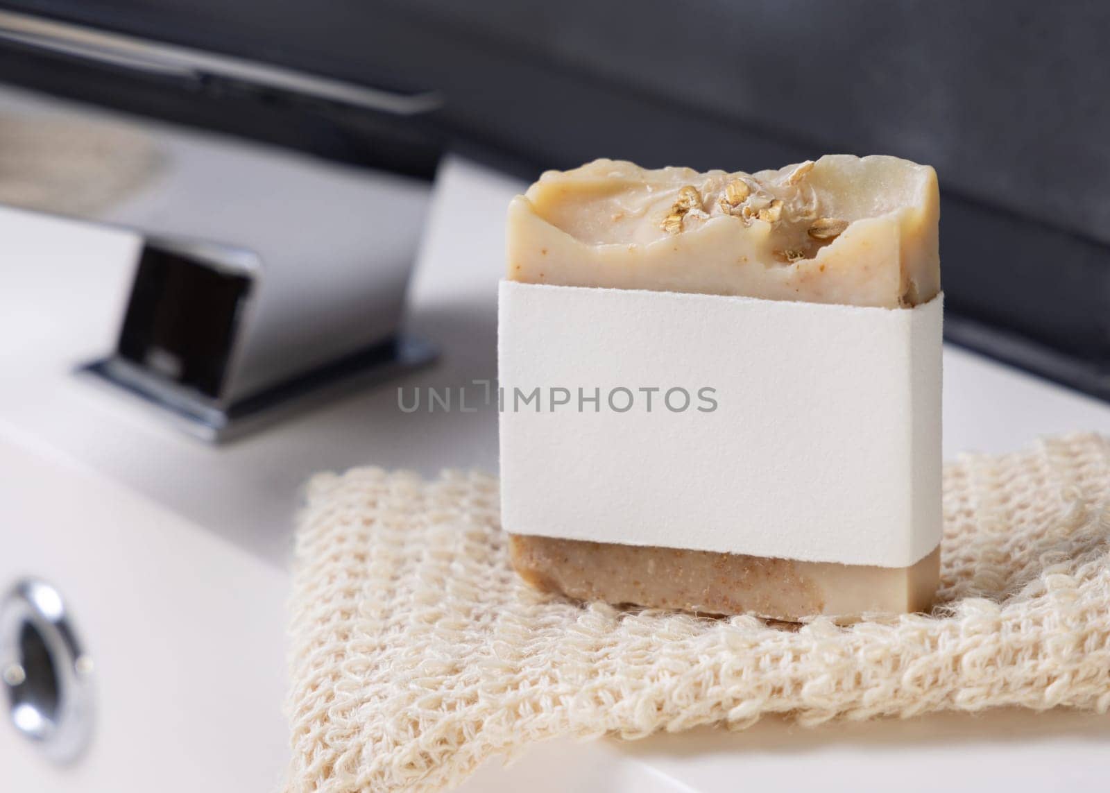 Beige handmade soap bar with blank label on sisal soap saver bag near modern faucet on basin in dark grey masculine bathroom close up, mockup. Natural herbal product for daily hygiene and bodycare