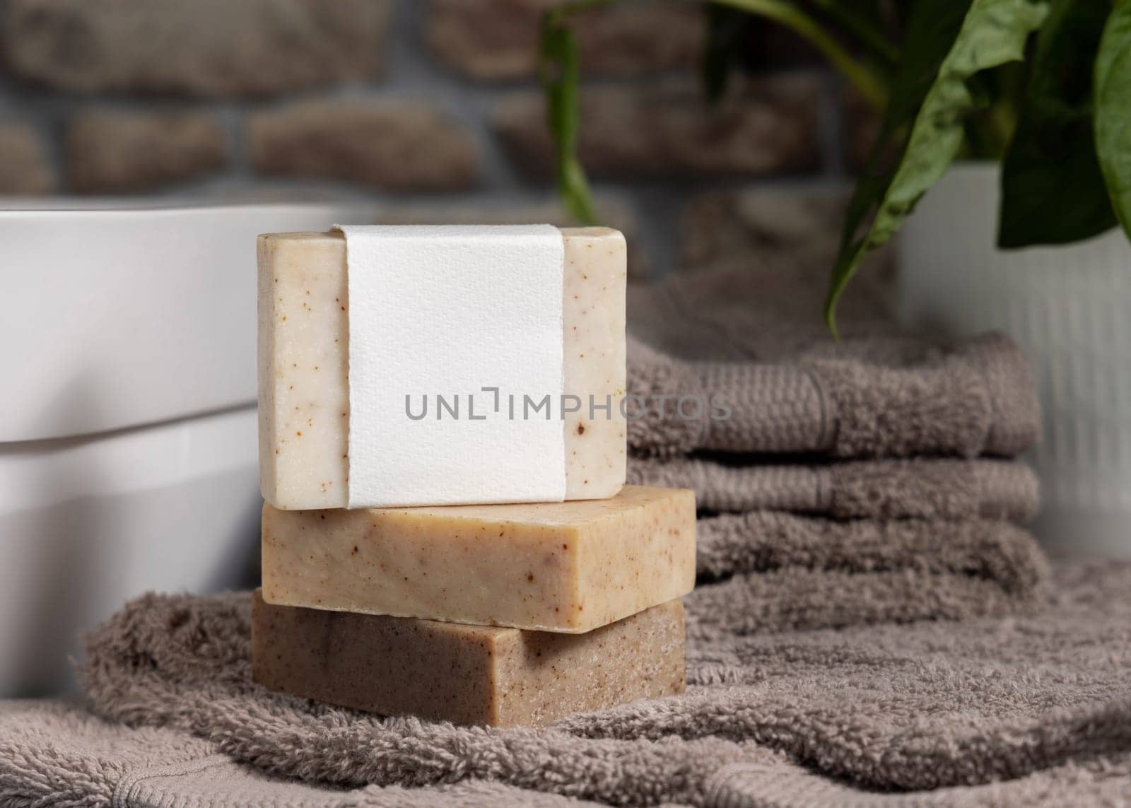 Soap bar with blank label on light brown folded towels, near green plant and vessel basin in bathroom, close up, brand packaging mockup. Men hygiene product
