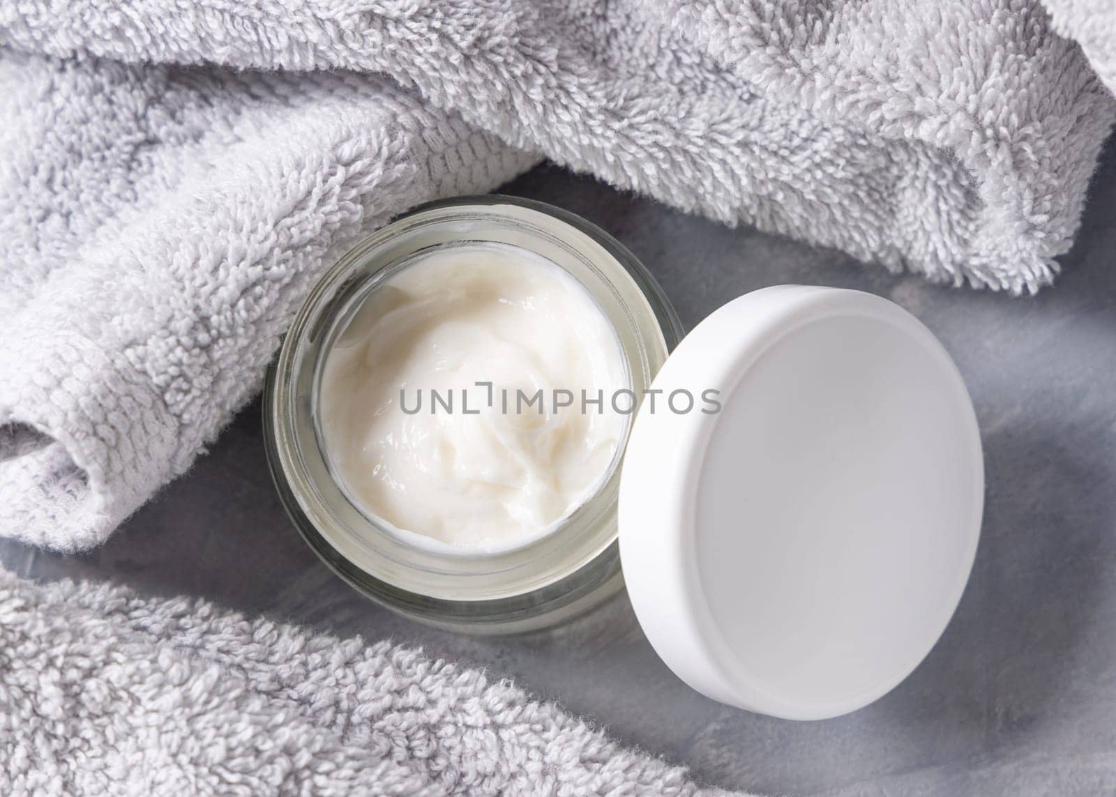White opened cream jar with blank lid near light grey towel in bathroom top view, brand packaging mockup. Natural organic cosmetics for daily facecare and spa procedures, copy space