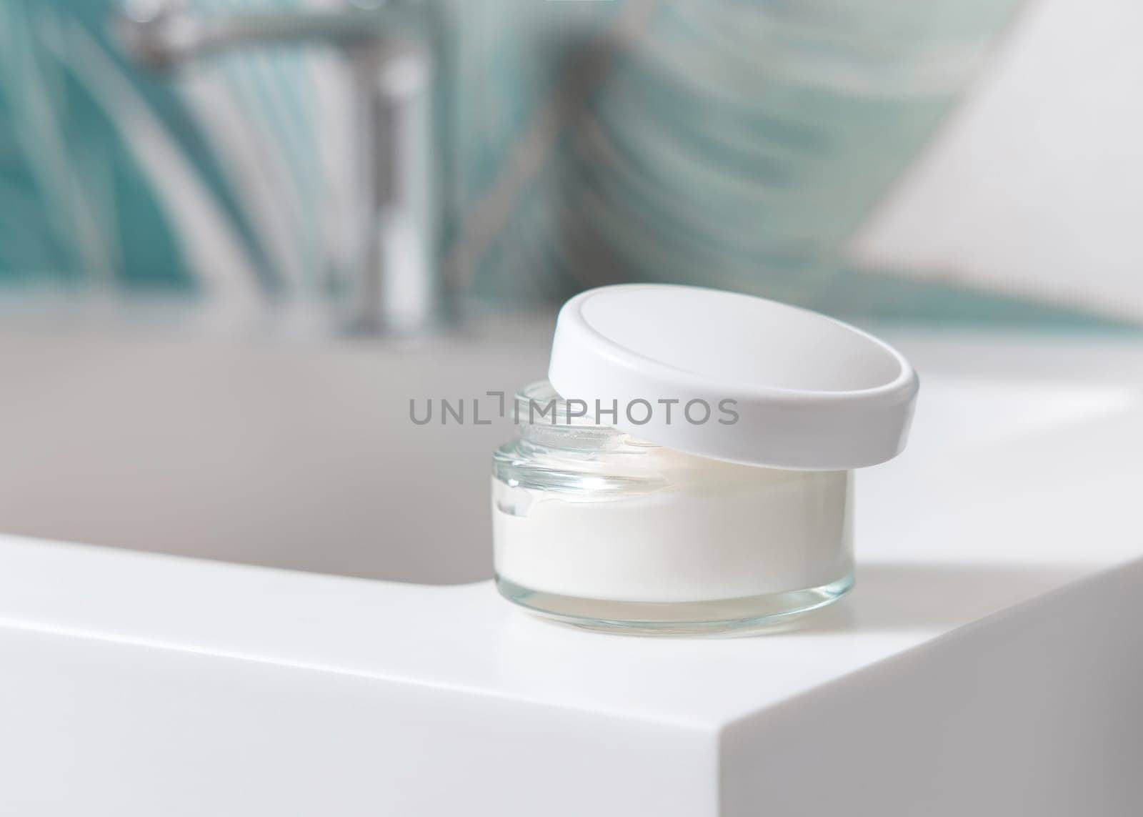 Opened cream jar with white blank lid on washbasin with faucet in green tropical bathroom close up, packaging mockup. Lifestile scene with ubranded natural facecare product