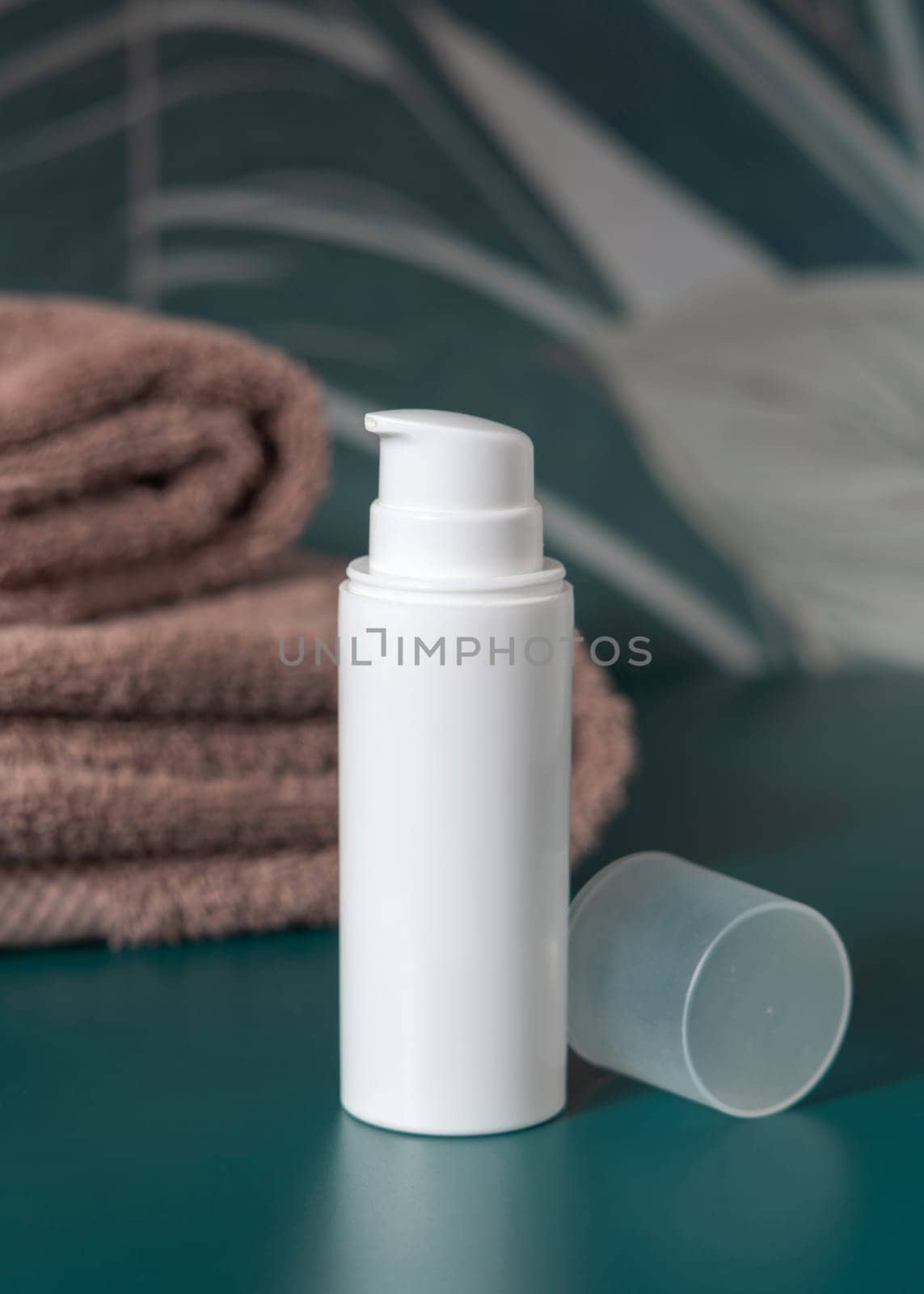 White cream one pump bottle near brown folded towels in dark green tropical bathroom close up, packaging mockup. Natural plant unbranded cosmetic container, copy space. Spa and wellness concept