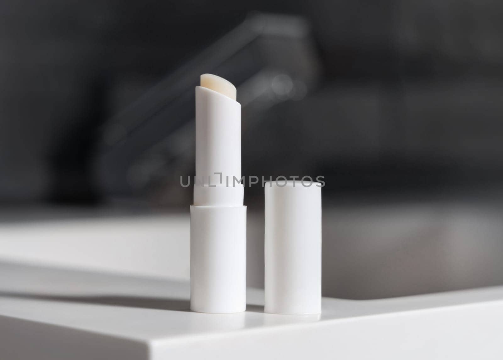 Opened lip balm stick with blank lid on basin with faucet in dark grey modern masculine bathroom close up, packaging mockup. Natural unbranded cosmetics for lipcare, copy space