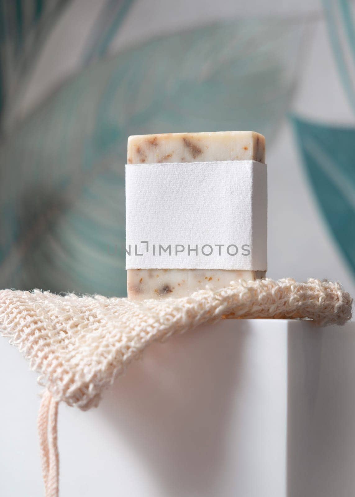 Beige handmade soap bar with blank label on sisal soap saver bag on washbasin in green tropical bathroom close up, mockup, copy space. Natural herbal unbranded product for daily hygiene