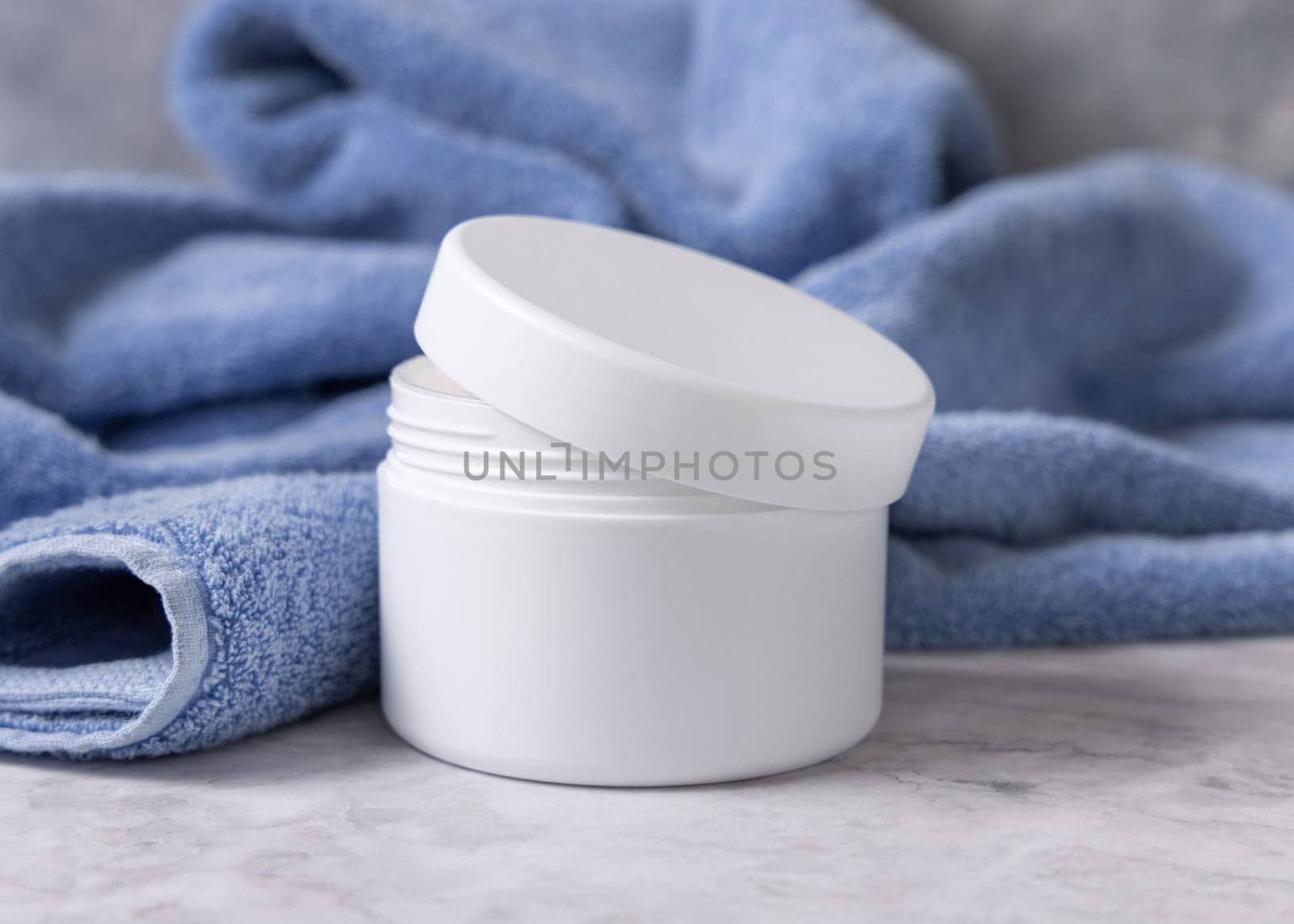 White opened cream jar with blank lid near blue bath towel close up, brand packaging mockup. Natural herbal organic cosmetics for daily facecare and spa procedures, copy space