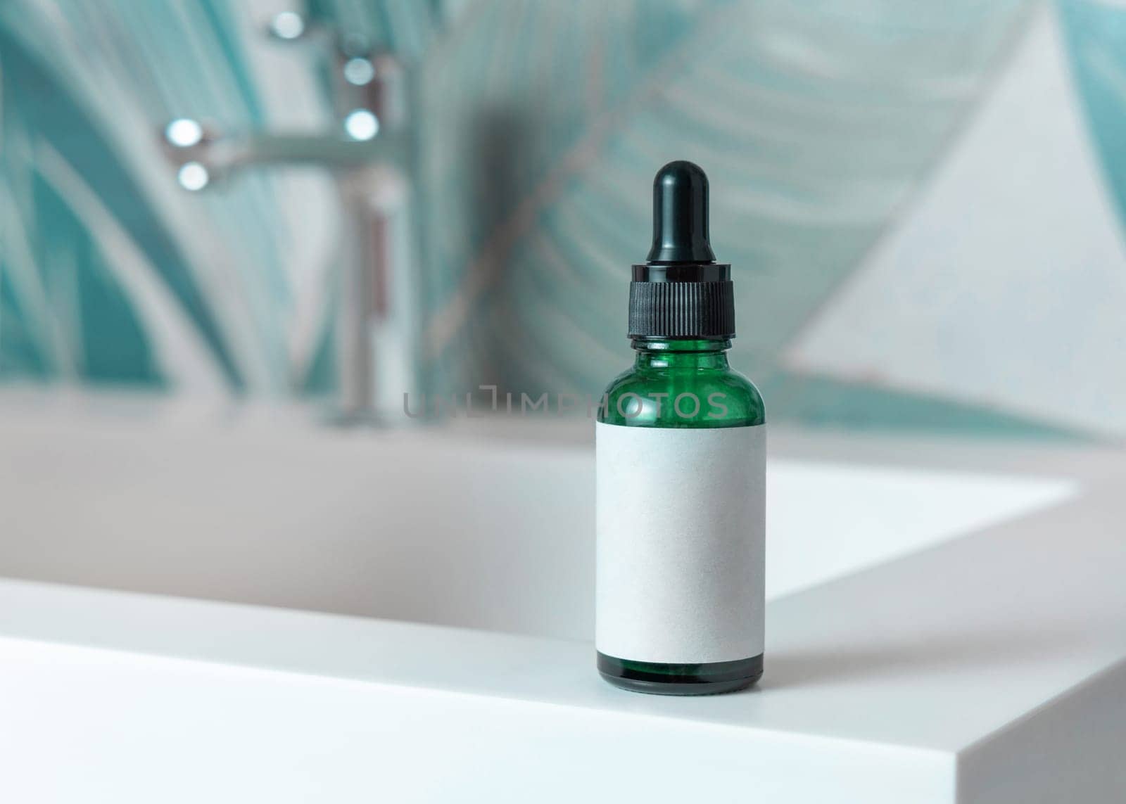 Green glass dropper bottle with pipette and blank label on washbasin with faucet in green tropical bathroom close up. Natural cosmetics - serum or essence. Daily beauty procedures
