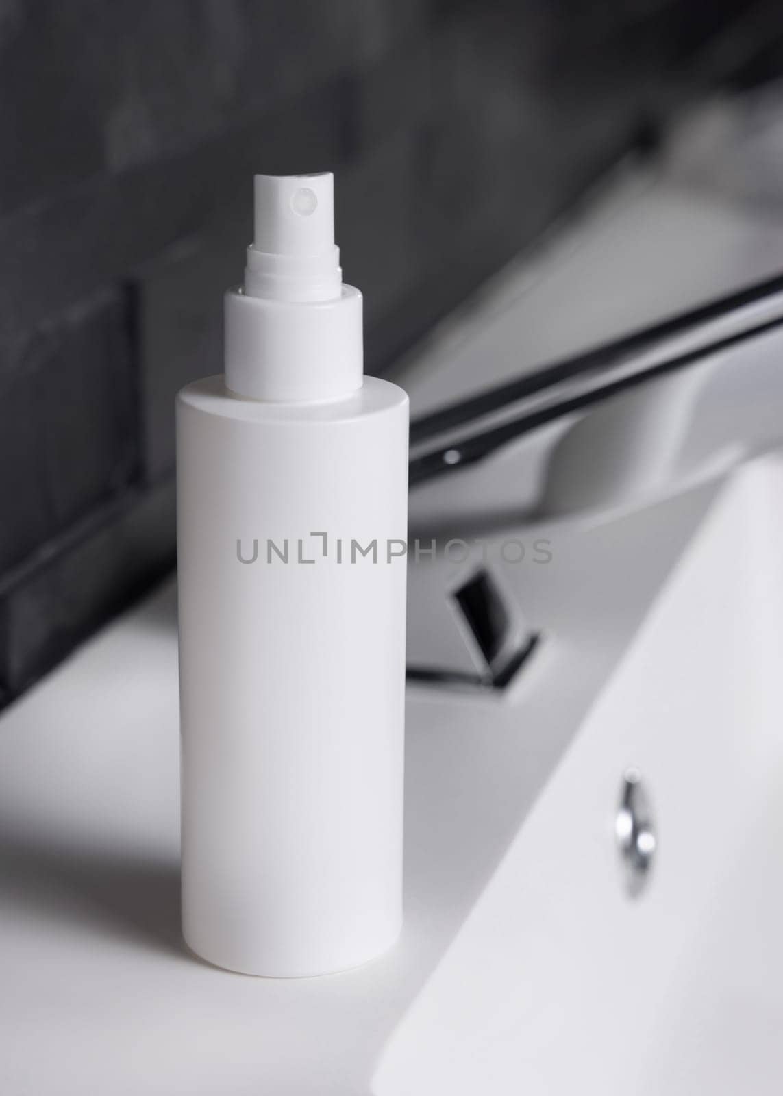 White spray bottle near modern faucet on basin in dark grey masculine bathroom close up, packaging mockup. Natural unbranded cosmetics with copy space. Spa and wellness concept