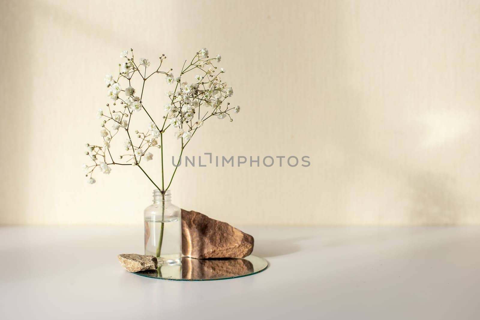 One branch of a white gypsophila flower standing on a little bottle and on a round mirror with stone on a beige background, copy space, abstract design concept. Mock up