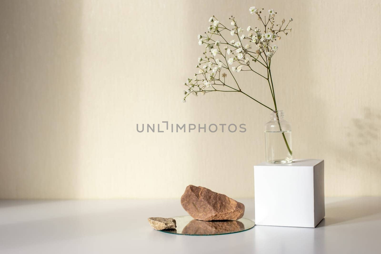One branch of a gypsophila flower standing on a white paper cube, near three stones on a round mirror, on a beige background, copy space, abstract design concept. Mock up