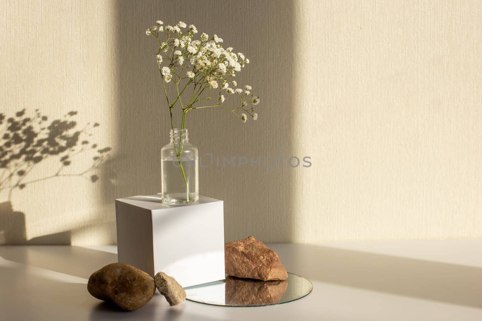 A branch of a gypsophila flower standing on a bottle on a white paper cube, near three stones on a round mirror, on a beige background, copy space, abstract design concept.