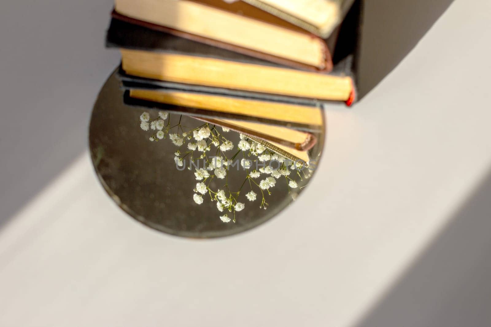 three old books lie with white gypsophila flowers and a round mirror on the table in the sunlight. Copy space. Top view