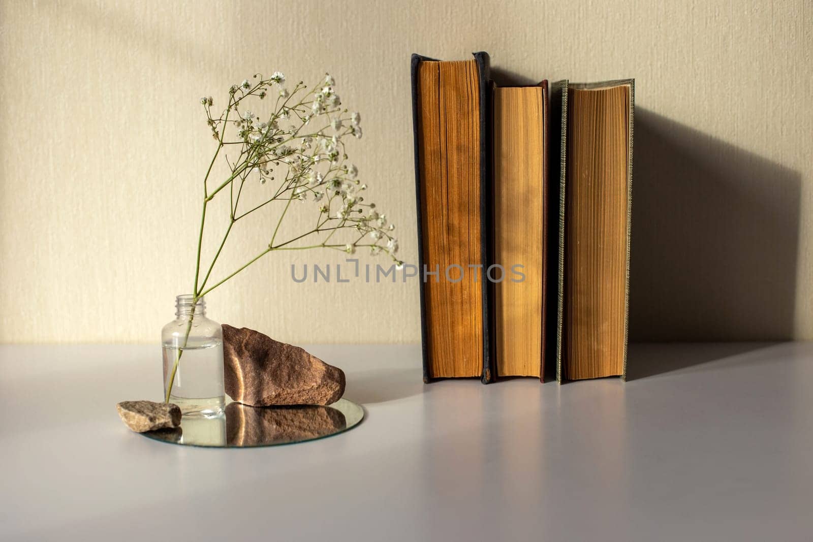 Three old books stand next to a branch of a white gypsophila flower standing in a small bottle and on a round mirror with stones, on a beige background, copy space, abstract design concept.