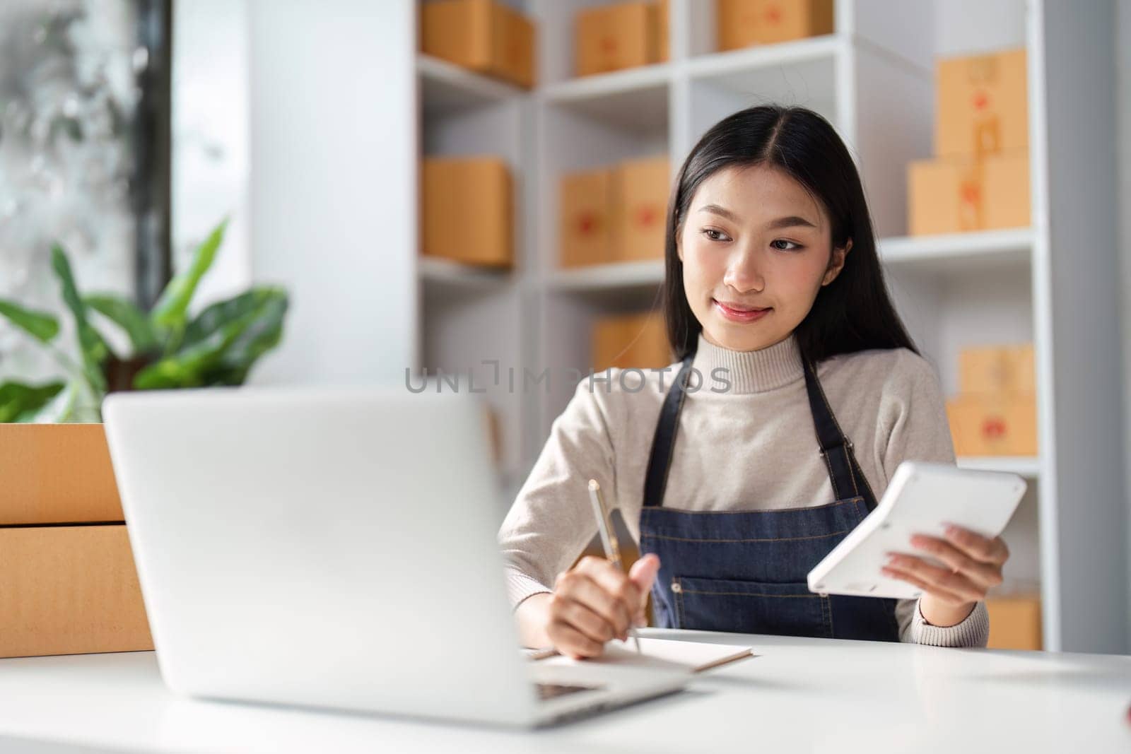 A young Asian woman examines e-commerce data on her tablet while taking notes, illustrating her analytical skills in a professional environment.