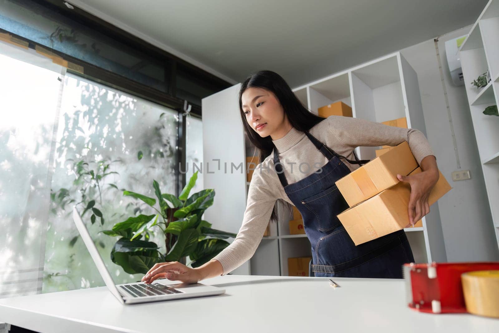 An e-commerce businesswoman managing online orders on her laptop while carrying packages in a modern office environment.