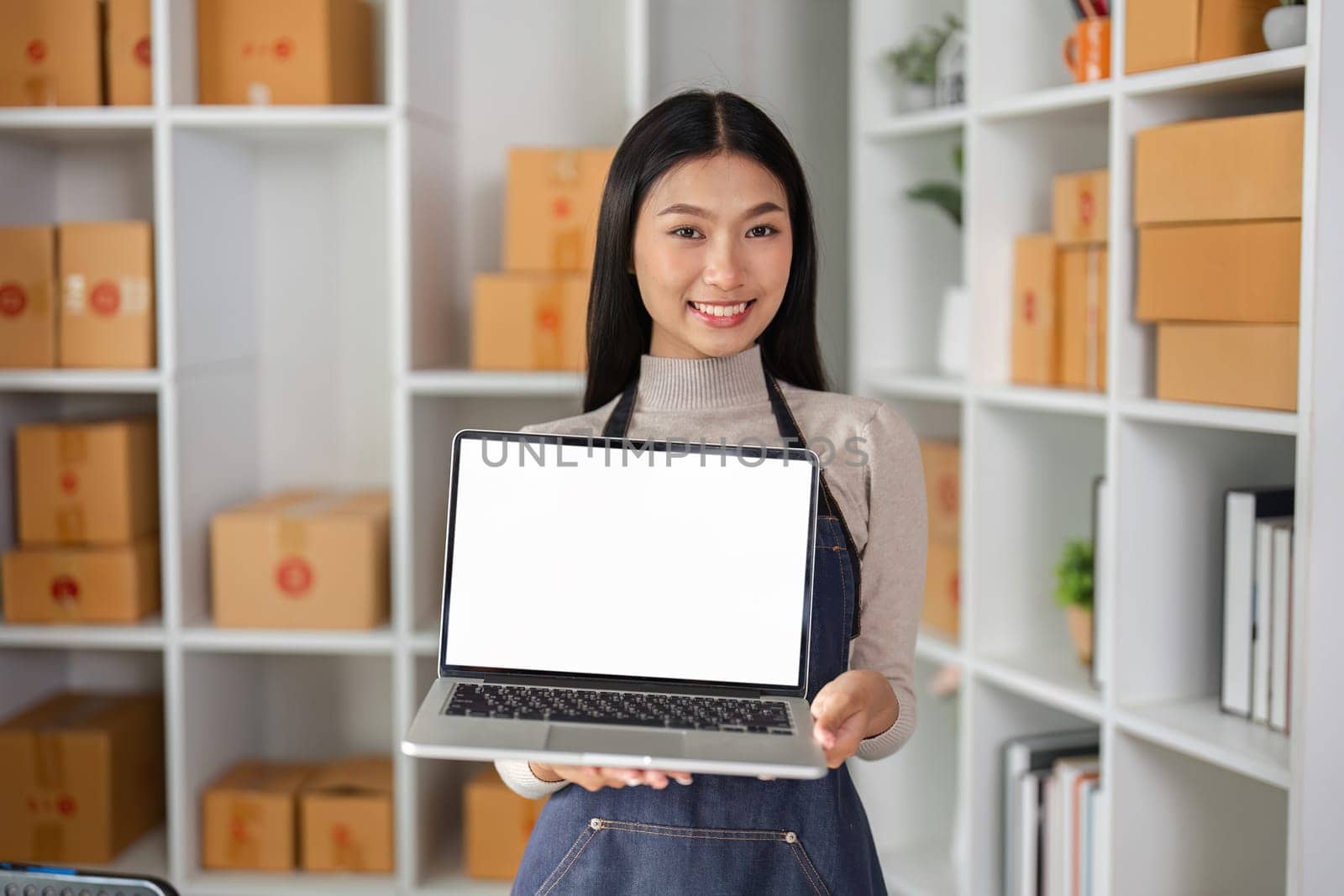 A smiling young female entrepreneur presents a laptop in a modern workspace, highlighting her business and creativity.