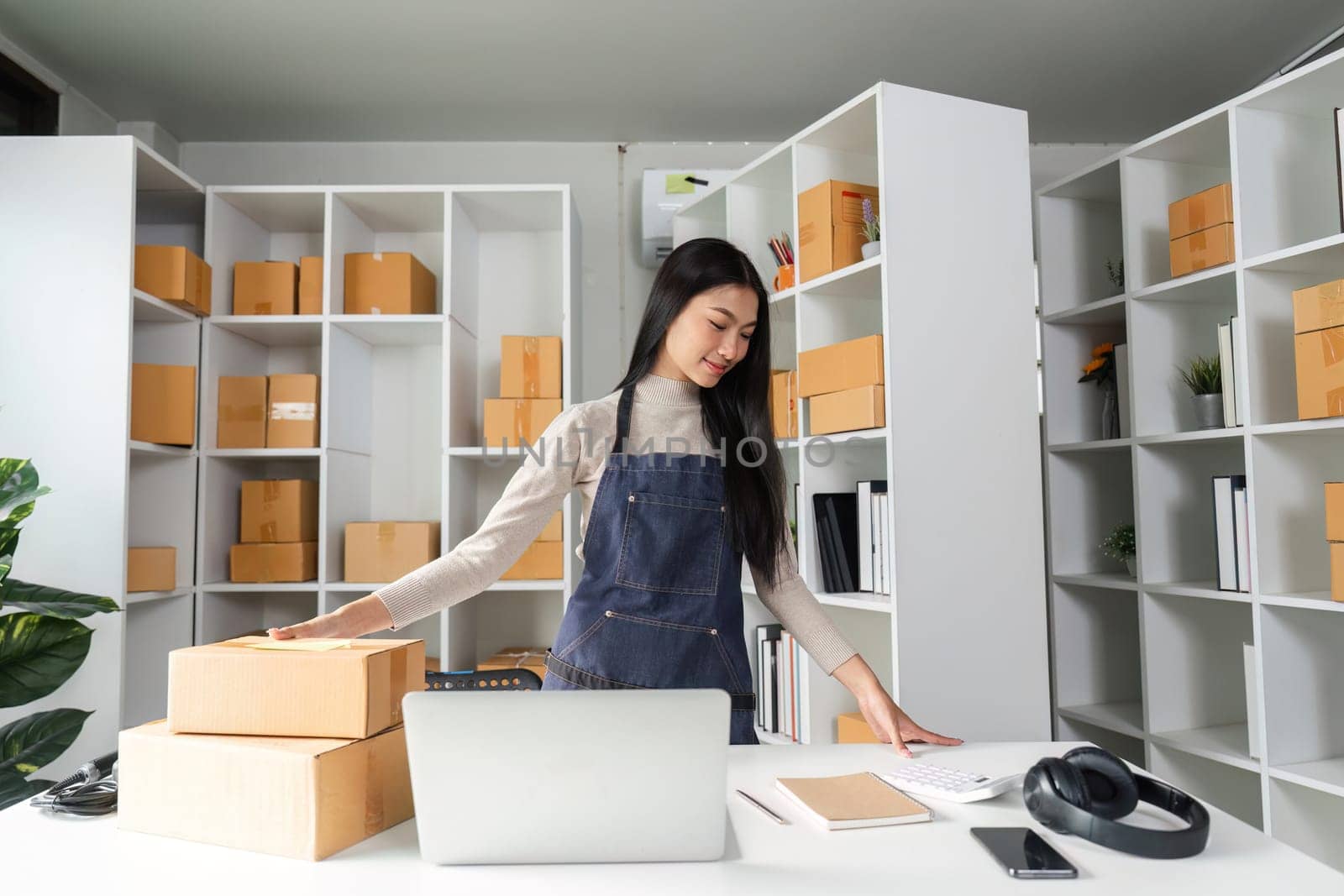 A focused young woman manages logistics in her stylish office, organizing packages while using her computer.