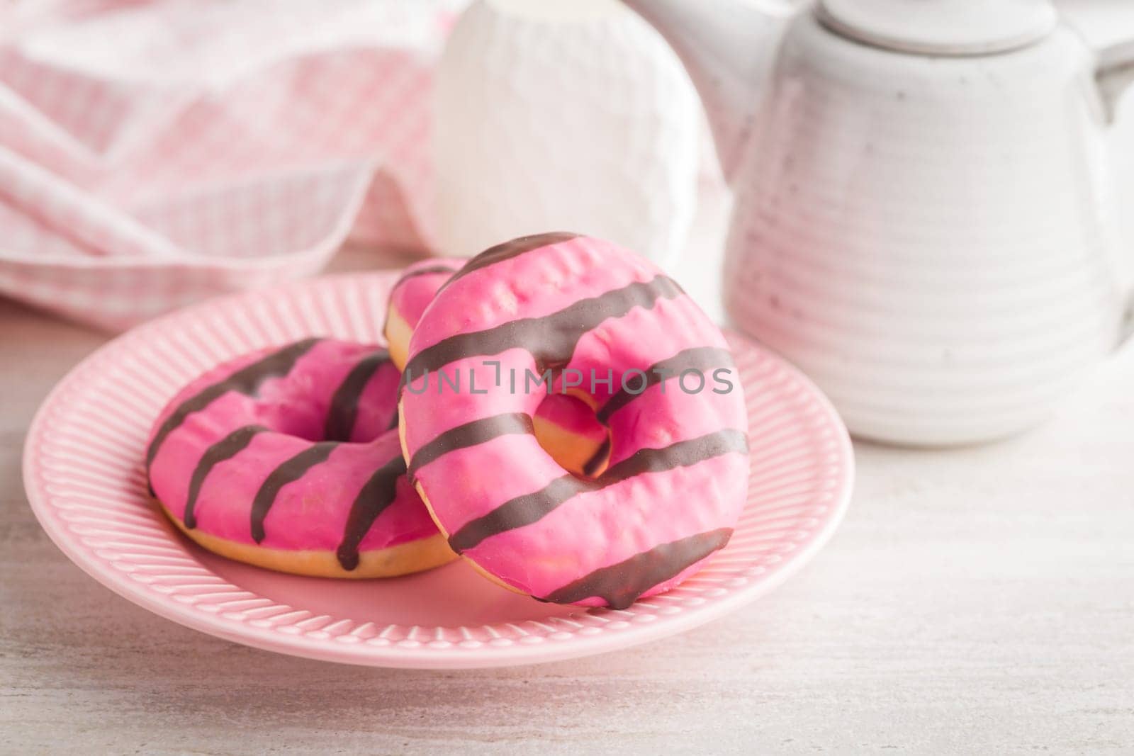 Sweet pink donut with chocolate strips on plate on a kitchen table.