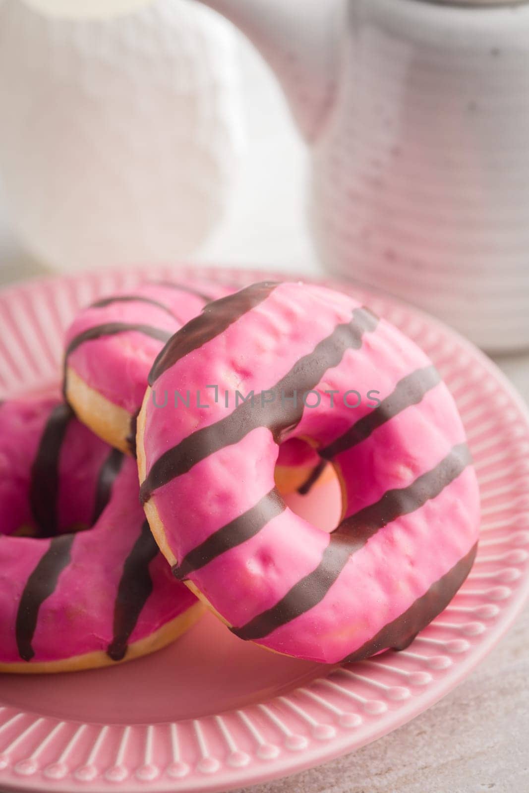 Sweet pink donut with chocolate strips on plate on a kitchen table.