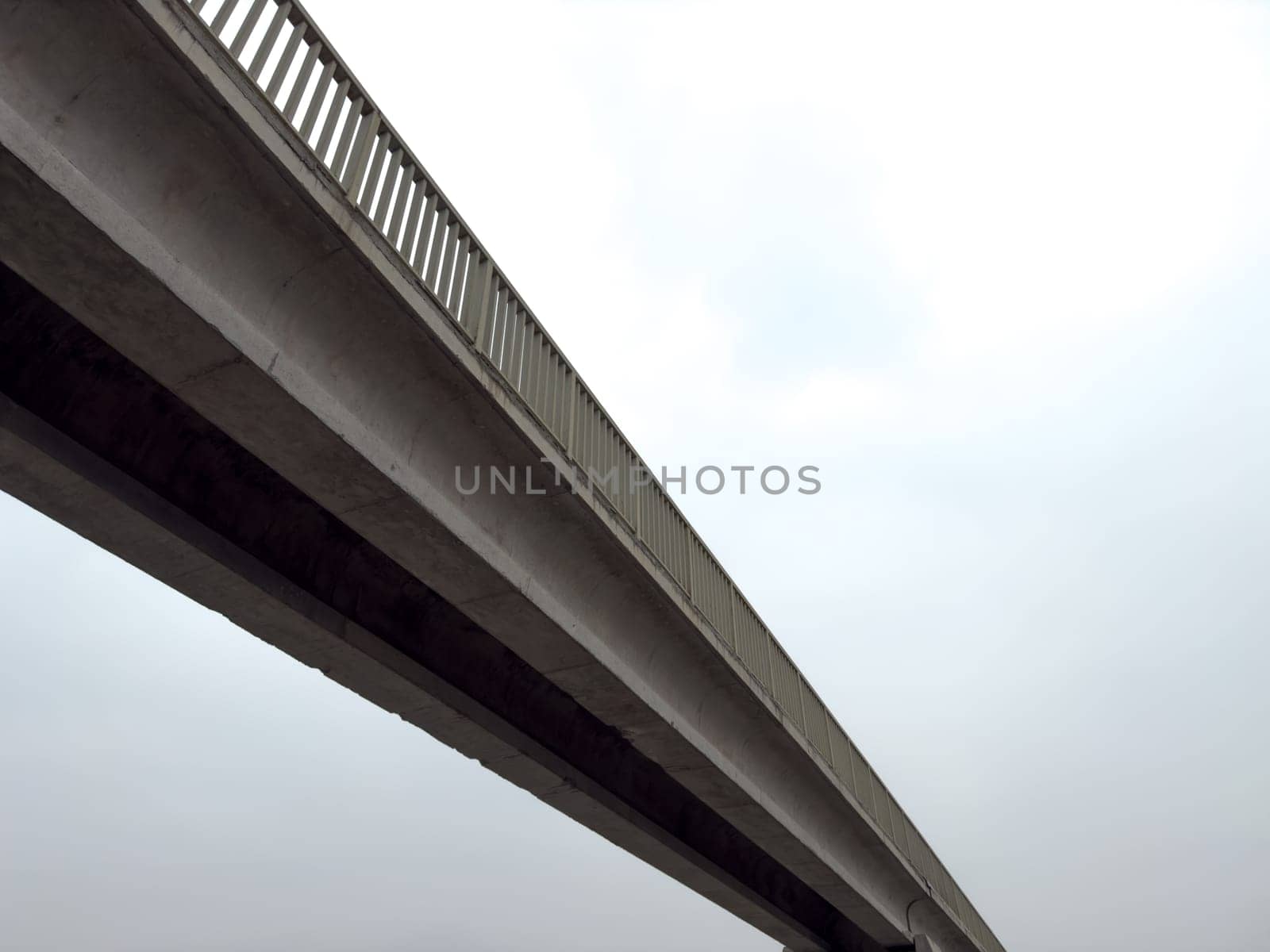 Long concrete bridge against dark sky. Cloudy day with footbridge across road for pedestrians. Simple concrete pedestrian bridge design made of concrete blocks and metal fencing for safety