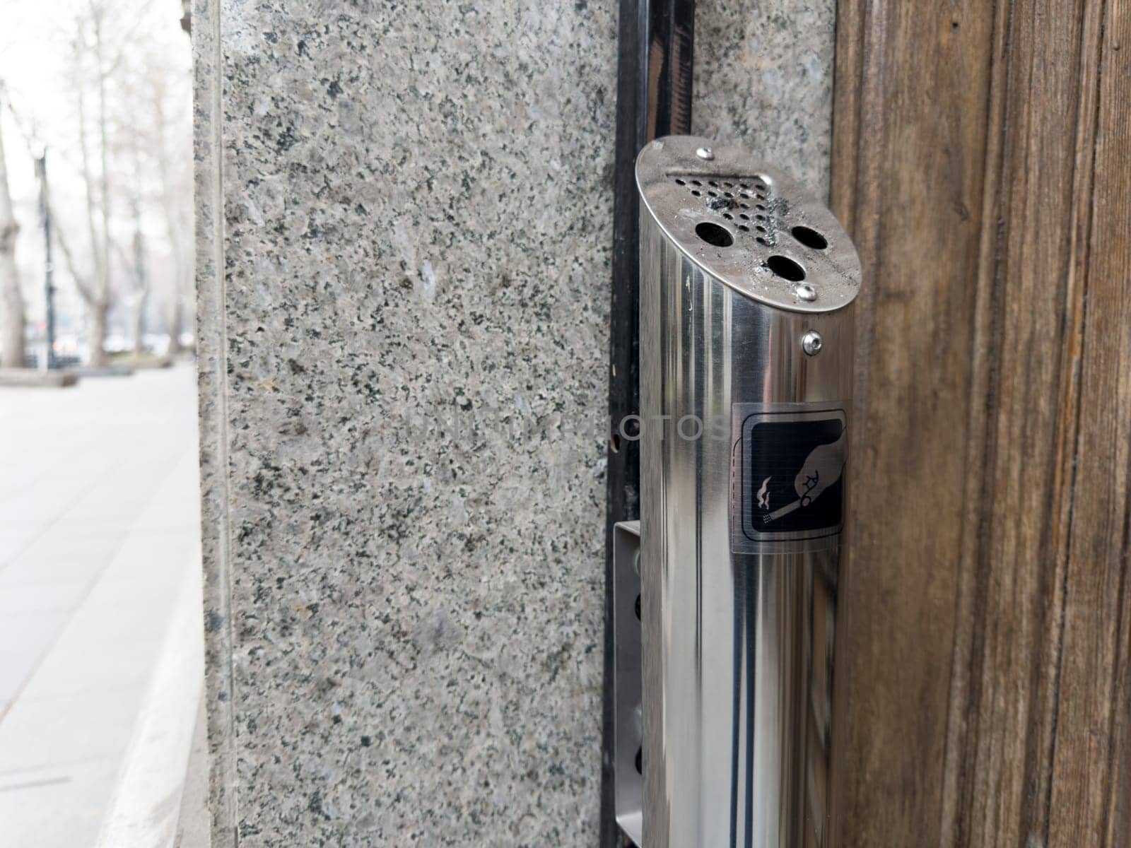Outdoor ashtray is attached to building for convenient smoking of cigarettes. Ashtray and trash can in smoking area. Bad habit of smoking cigarettes. Steel trash can post at entrance to building