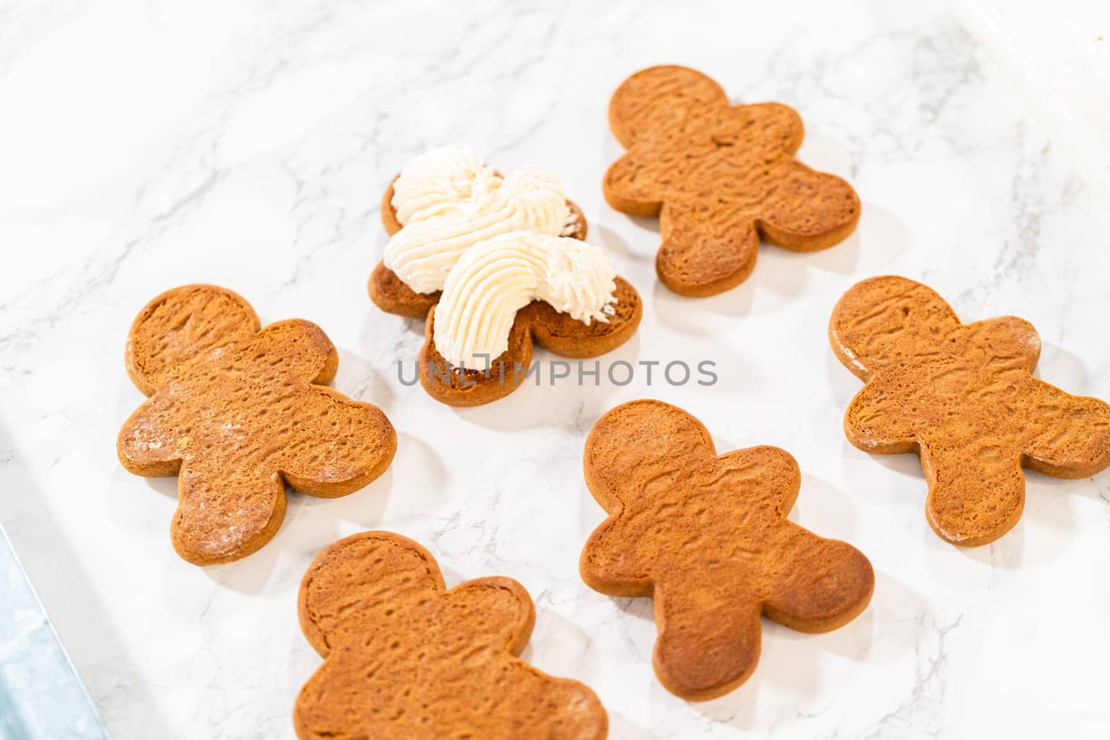 Gingerbread cookies await their second halves on a marble surface, each meticulously piped with buttercream to craft delightful sandwich treats. The precision of the piping adds a festive touch.