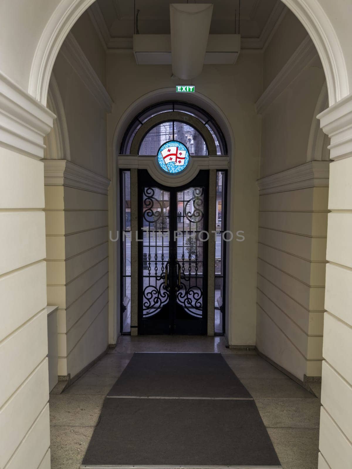 The exit from building with beautiful arch and white walls with large black doors with glass windows. Georgian flag at the exit of building. Beautiful passage to doors in Georgian building