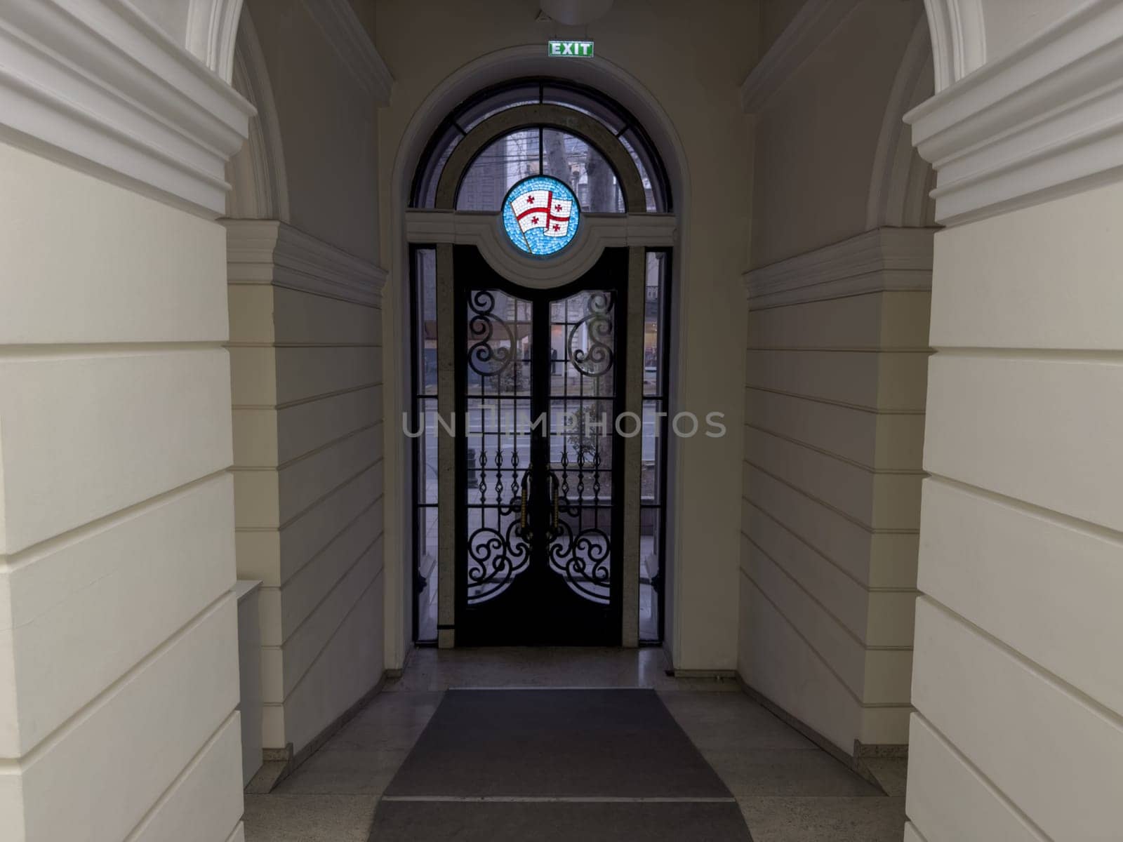 Corridor to exit door in Georgian building with symbol of country above large black doors with glass windows. Georgian flag at exit of building through arch and large doors
