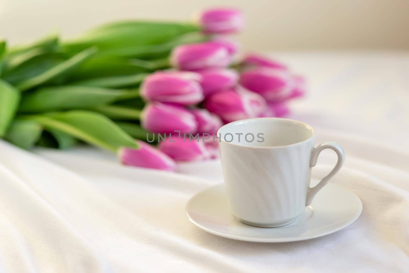 A white cup with a saucer and a bouquet of beautiful pink tulips lies on a white tablecloth. close up. copy space