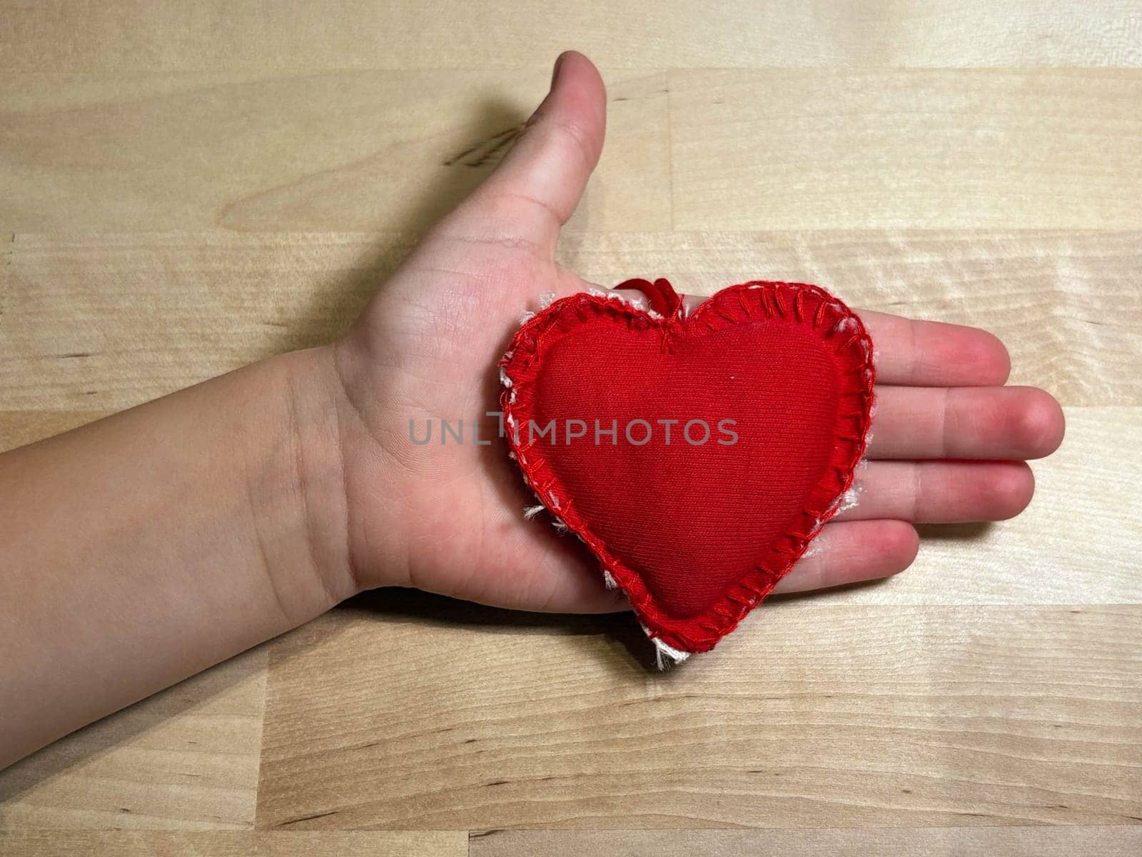 Heart sewn from red fabric in the hand of a small child on a wooden background. Love