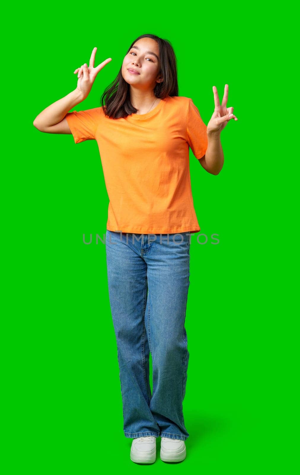A young woman is standing confidently with both hands forming peace signs. She wears an orange shirt and blue jeans, showcasing a joyful expression.