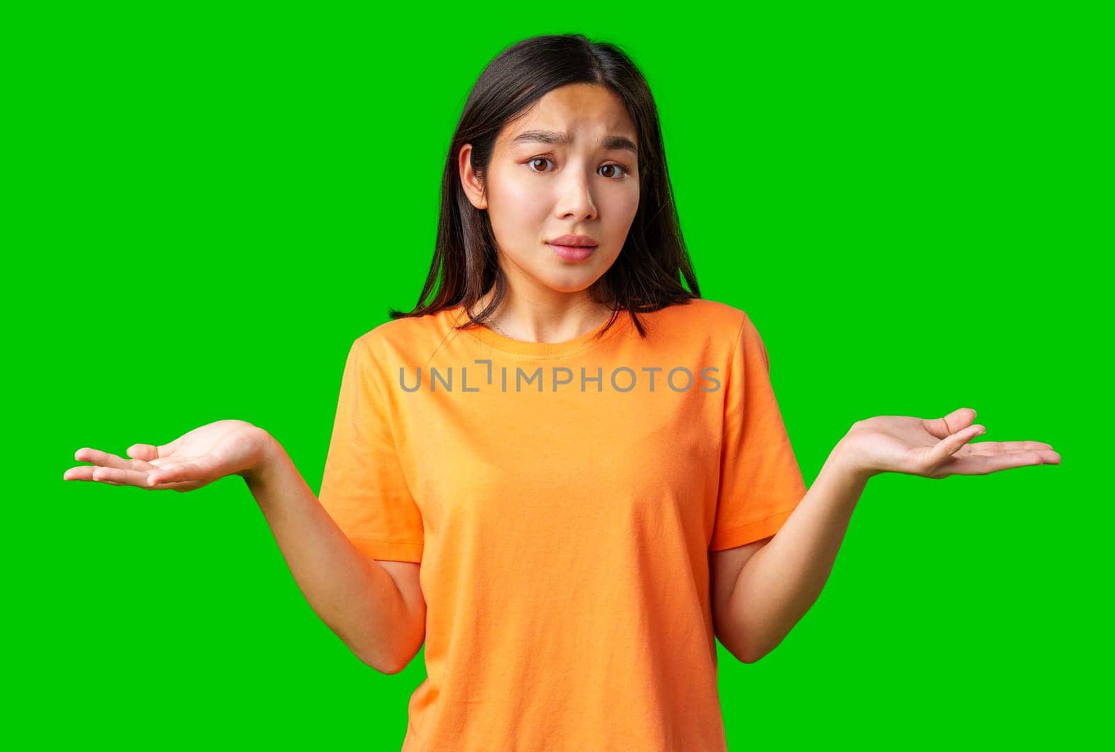 A young woman stands against a green background, wearing an orange shirt, raising her hands in a gesture of confusion or uncertainty, as if contemplating her next move or decision.