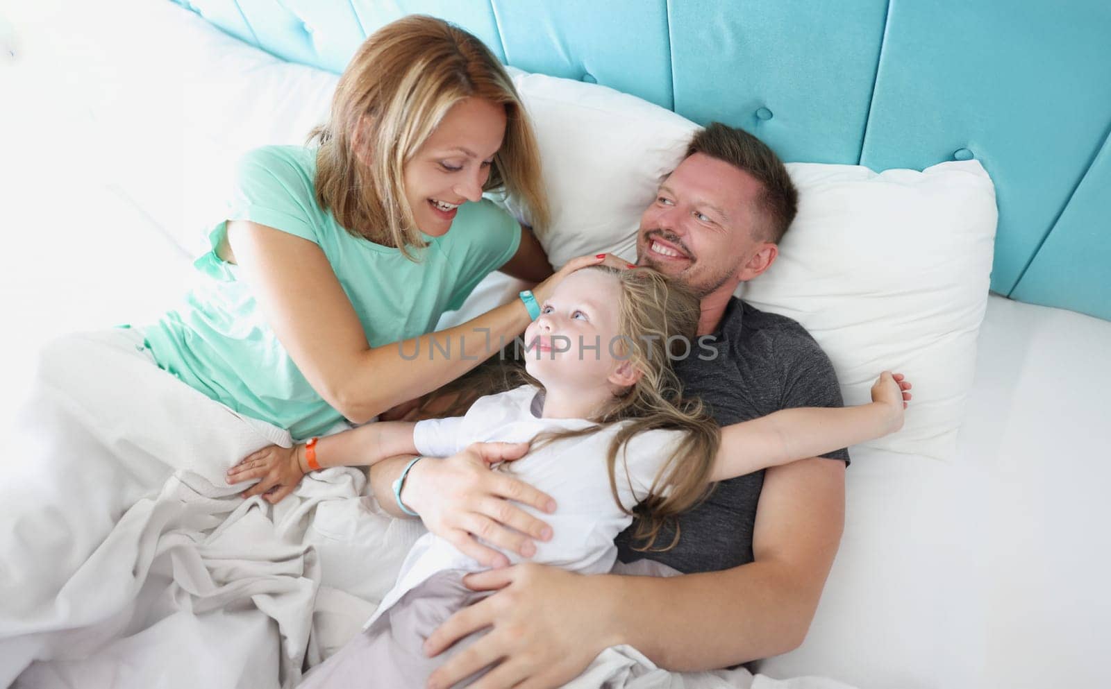 Parents and child lie in bed laughing and hugging. by kuprevich