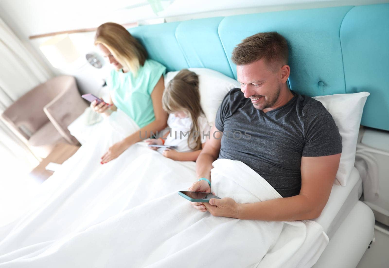 Parents and child lie in bed, each holding smartphone in his hands. Uncontrolled use of modern gadgets by families concept