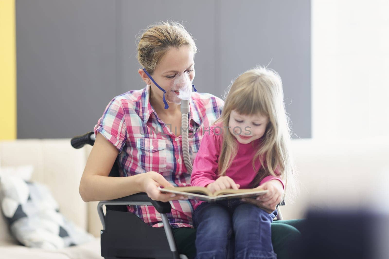 Woman with an oxygen mask and in wheelchair is reading book with little girl by kuprevich